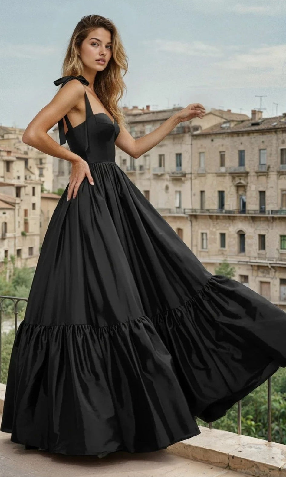 A woman wears the PromGirl Exclusive Prom Dress with Shoulder Bows in black, standing on a balcony with historic stone buildings in the background.